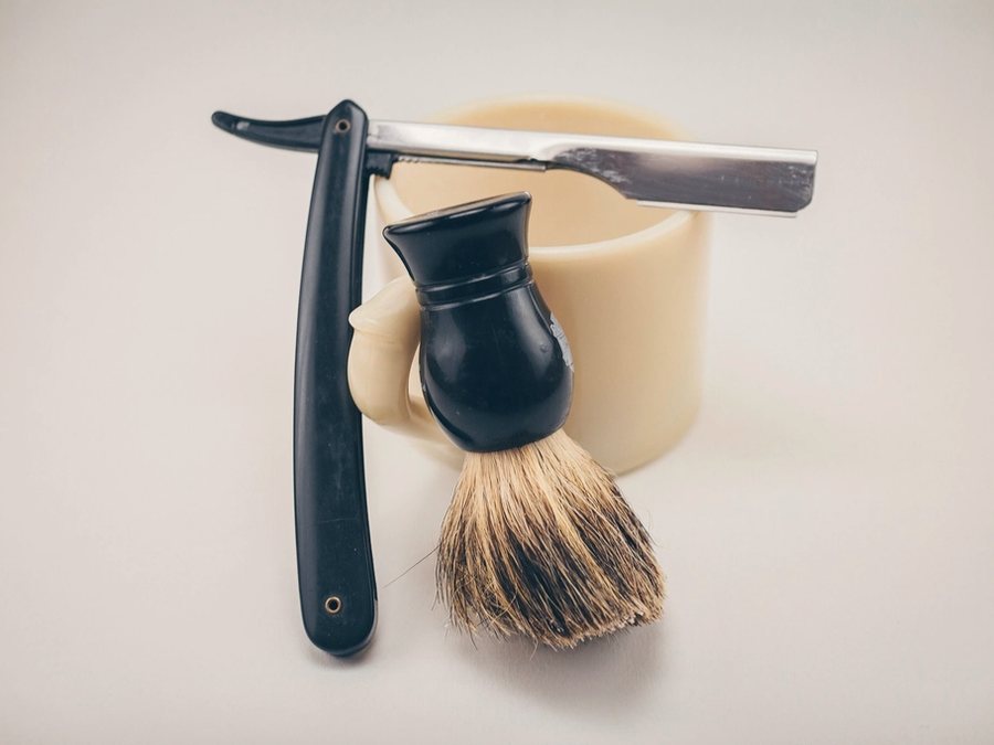 A straight razor and a badger shaving brush resting beside a bowl of lather.